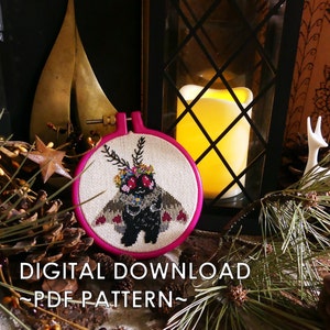 Puede incluir: Un aro de bordado rosa con tela blanca en su interior. La tela tiene un diseño de punto de cruz de una polilla con una corona de flores y alas en forma de corazón. El texto "DIGITAL DOWNLOAD ~PDF PATTERN~" está debajo del aro.