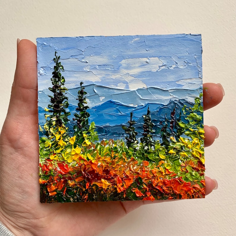 Op de afbeelding: Een klein schilderij van een berglandschap met een blauwe lucht, groene bomen en oranje en gele bloemen op de voorgrond.