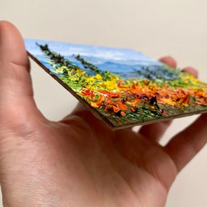 Op de afbeelding: Een klein schilderij op een houten paneel dat een landschap voorstelt met een blauwe lucht, groene bomen en een veld met gele en oranje bloemen.