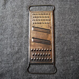 May include: Vintage metal cheese grater with a rectangular design and a handle. The grater has numerous small holes and two angled blades. The text "BROMCO MADE IN U.S.A." is visible on the grater.