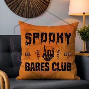 May include: Orange Halloween-themed pillow with the words "Spooky Babes Club" in black, along with skeleton graphics and the year 1629. The pillow is square and sits on a gray chair.