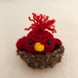 May include: A red crocheted bird with a yellow beak and black eyes sits in a brown crocheted nest.
