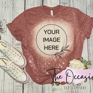 May include: A dusty rose colored t-shirt with a bleached circle in the center. The circle has the text "YOUR IMAGE HERE" inside. The t-shirt is laying on a white wooden surface with a pair of white sneakers and a bouquet of white flowers.