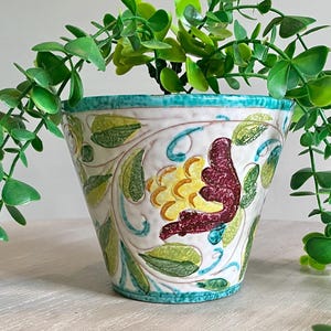 May include: A ceramic planter with a white base, featuring colourful floral and bird designs. The planter has a turquoise rim and is filled with green, leafy plants. The arrangement is set on a light-coloured surface.