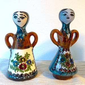 May include: Two ceramic figurines with a folk art design. Each figurine has a stylized face, brown handles, and a skirt decorated with floral patterns in blue, red, and yellow. The figurines are approximately 15 cm tall.