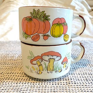 May include: Two stacked white ceramic mugs with brown trim and handles. The top mug features illustrations of tomatoes and a pumpkin. The bottom mug displays mushroom illustrations. The mugs are on a white and patterned surface.