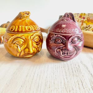 May include: Two ceramic jars, one for onions and one for beetroot, each with a face design. The onion jar is golden brown with "ONIONS" on top. The beetroot jar is dark red with "BEETROOT" on top. Both have a whimsical, anthropomorphic design.