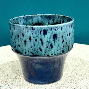 May include: A two-tone ceramic plant pot. The upper section is blue-green with a speckled pattern, while the base is dark blue. The pot has a cylindrical top and a flared base. The speckled design gives the pot a unique, artistic appearance. Suitable for indoor plants.