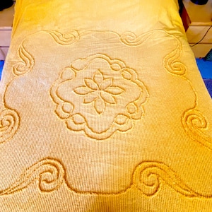 May include: A mustard yellow bedspread featuring a textured, ribbed surface. The centre displays a raised, tufted floral motif enclosed within a scalloped border, with additional swirling, scroll-like patterns positioned at each corner.