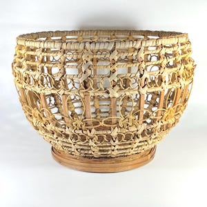 May include: A hand-woven basket with a wide opening and a rounded base. The basket is made from natural reeds and features an intricate open weave pattern. The basket rests on a circular base.