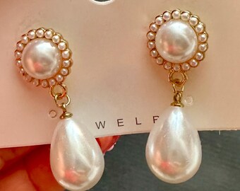 May include: A pair of elegant earrings featuring a large, round, white pearl surrounded by smaller pearls in a gold-tone setting. Each earring has a teardrop-shaped pearl pendant. The earrings are on a white card.