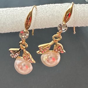 May include: A pair of gold-toned drop earrings. Each earring features a pearlescent, teardrop-shaped bead, accented with pink crystals and a clear crystal. The earrings have a leaf-like design.