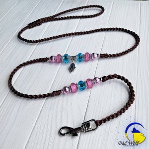May include: A brown braided dog leash with a silver clasp and pink and blue crystal beads. The beads have a silver charm in the shape of a hand with an eye in the center.