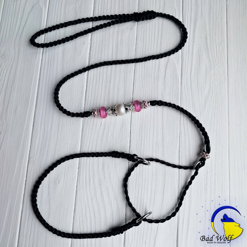 May include: Black braided dog leash with a pink and silver beaded charm. The leash is made of two separate pieces, a longer leash and a shorter loop.