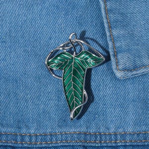 Op de afbeelding: Zilverkleurige blad-vormige broche met een levendige groene emaille afwerking. De broche heeft ingewikkelde details, waaronder adering op het blad en een wervelend zilveren frame. De broche is geplaatst tegen een denim achtergrond.