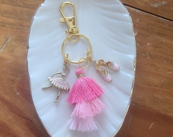 Puede incluir: Un llavero dorado con un borla rosa y blanca, un amuleto de bailarina y un par de zapatillas de ballet rosas.