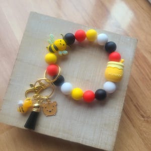 Puede incluir: Un llavero de cuentas de colores con un tema de Winnie the Pooh. El llavero presenta una borla negra, un dije de abeja y un dije de Winnie the Pooh. La pulsera está hecha de cuentas redondas en amarillo, rojo, negro y blanco. También se incluye un dije de tarro de miel.