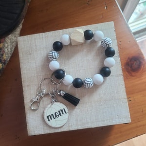 May include: A white and black beaded bracelet with volleyball charms and a wooden bead. A keychain with a volleyball charm and the word "mom" is attached to the bracelet.