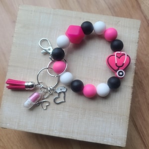 May include: A pink, black, and white silicone beaded bracelet with a heart charm featuring a stethoscope. The bracelet has a silver key ring with a small pink tassel, a pill charm, and a nurse charm.
