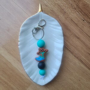 May include: A keychain featuring a charm in the shape of a cowboy boot with turquoise and brown accents. The boot is attached to a silver keyring with turquoise and black beads. The keychain is resting on a white leaf-shaped dish.