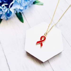 May include: A gold chain necklace with a red enamel ribbon pendant. The necklace is on a white hexagonal display stand.