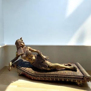 May include: A gold-toned statue of a woman reclining on a decorative, ornate, patterned cushion. The woman is wearing a gold headdress and jewelry.