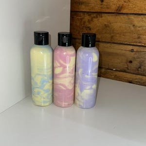 May include: Three bottles of lotion with swirl patterns in shades of blue, pink, and purple. The bottles have black lids and are arranged in a row on a white surface.
