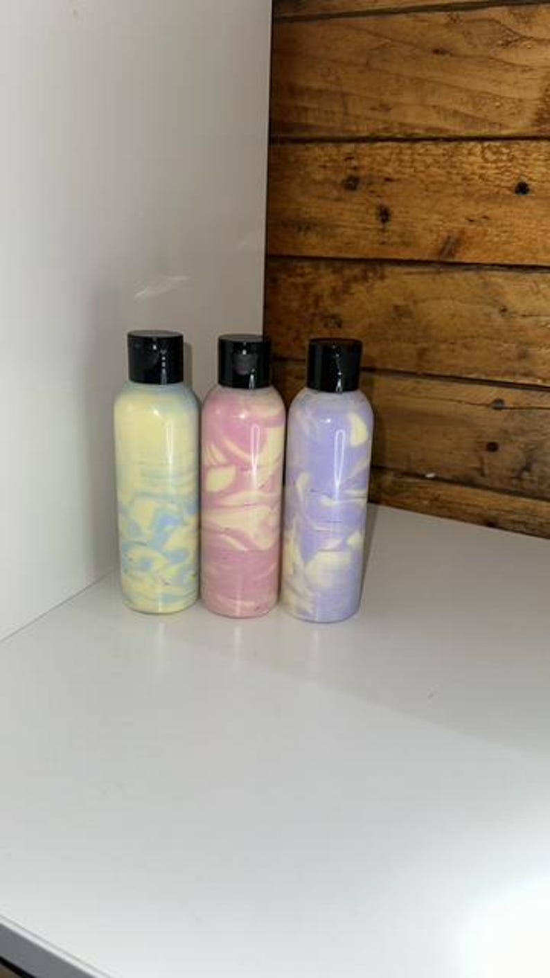 May include: Three bottles of lotion with swirl patterns in shades of blue, pink, and purple. The bottles have black lids and are arranged in a row on a white surface.