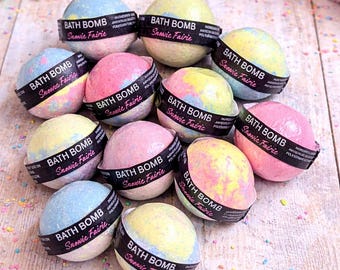 May include: An assortment of colorful bath bombs with the text "BATH BOMB Snowie Fairie" on a black band. The bath bombs are in shades of blue, pink, and yellow. The image includes flowers and a pink towel.