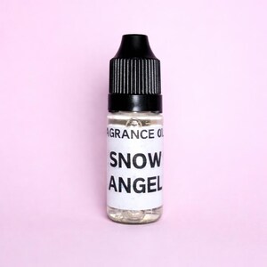 May include: A small glass bottle of fragrance oil with a black cap. The label reads "SNOW ANGEL" in bold white letters. The bottle is filled with a clear liquid and set against a pink background.