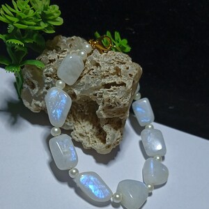 May include: A necklace featuring rectangular, iridescent moonstone beads interspersed with small, round pearls. The beads have a milky white base with flashes of blue and purple. The necklace is secured with a gold clasp.