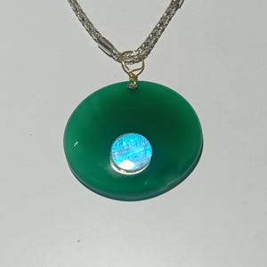 May include: A green circular pendant with a blue gemstone center, suspended from a silver chain. The pendant is a vibrant green, and the gemstone reflects a range of blue hues. The chain is a delicate silver weave.