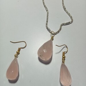 May include: A jewelry set featuring a silver chain necklace with a teardrop-shaped pink gemstone pendant. Matching earrings with gold-colored hooks and teardrop-shaped pink gemstones are also included. The set is displayed on a white surface.