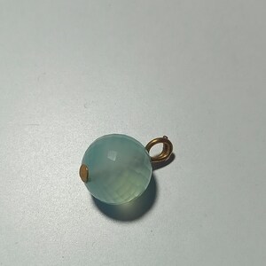 May include: A faceted, light blue gemstone pendant with a gold-colored bail. The round bead has a subtle gradient of color, transitioning from a pale green at the base to a light blue at the top. A small, gold-colored accent is on the side.