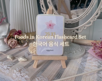 May include: A flashcard from a Korean food set, displayed on a small wooden easel. The card features a pink flower graphic and Korean text. The text "Foods in Korean Flashcard Set" is above the Korean text. The word "eggplant" is at the bottom.