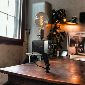 Könnte beinhalten: Eine Vintage-Kamera-Lampe mit einer klaren Glühbirne aus Glas und einem spiralförmigen Filament. Die Kamera ist schwarz und silber mit einem Stativ.