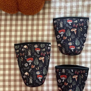 May include: Three black fabric cozies with a woodland pattern of mushrooms, flowers, and insects. The cozies have a black trim and are arranged on a brown and white checkered surface. A fuzzy orange pillow is in the background.
