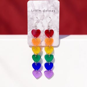 Puede incluir: Pendientes de corazón con los colores del arcoíris y ganchos plateados. Cada pendiente presenta siete dijes en forma de corazón en rojo, naranja, amarillo, verde, azul y morado. Se exhiben en una tarjeta blanca con el texto "Little.galaxy".