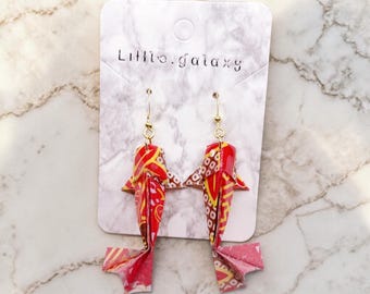 May include: A pair of origami koi fish earrings. The earrings are red, yellow, and white with gold-colored ear wires. The earrings are displayed on a white card with the text "Little.galaxy".