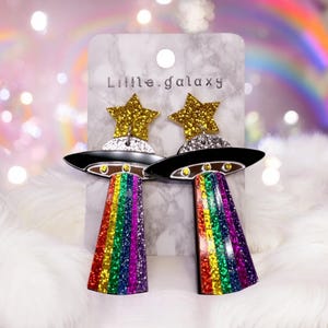 May include: A pair of black and silver UFO earrings with rainbow glitter stripes. The earrings feature a gold glitter star on top of each UFO.