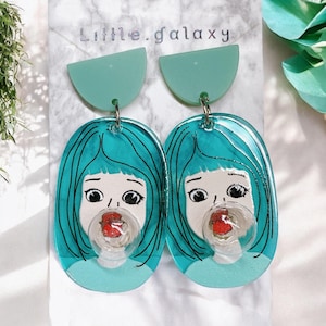 Könnte beinhalten: Ein Paar türkisblaue Ohrringe mit einem verspielten Design eines Mädchens, das eine Blase bläst. Die Ohrringe sind aus Harz gefertigt und haben ein halbrundes Oberteil.