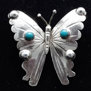 May include: A silver butterfly brooch with turquoise gemstone accents. The brooch features detailed wing patterns and rounded silver accents. The butterfly's body and antennae are also silver, creating a striking contrast against the black background.