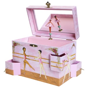 May include: Pink wooden ballerina music box with a mirror inside the lid. The box features a ballerina figurine that spins when the music plays. The box has two small drawers on the sides and a larger drawer on the front. The box is decorated with a floral design and a ballerina theme.