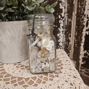 May include: A clear glass jar with a rusted metal wire handle filled with a collection of assorted white, cream, and beige buttons of various shapes and sizes, including round, flat, and bow-shaped designs, visible through the transparent glass.