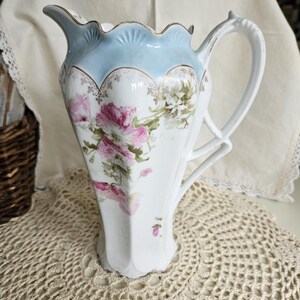 May include: A white porcelain pitcher featuring a scalloped rim with a light blue band, gold trim, and a floral pattern of pink and white flowers. The pitcher has a curved handle and a molded, fluted body with a decorative spout and base.