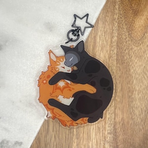 Puede incluir: Un llavero acrílico transparente con dos gatos abrazándose. Uno es naranja con detalles blancos, el otro es negro con detalles grises. Un cierre negro en forma de estrella está unido en la parte superior. Los gatos se abrazan.