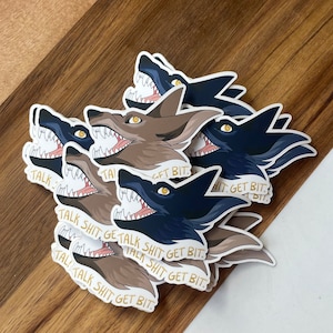 May include: A collection of stickers featuring a snarling wolf illustration in shades of brown and navy blue. The stickers have the text "TALK SHIT GET BIT" in gold lettering. The stickers are on a wooden surface.