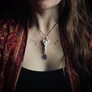 May include: A distinctive pendant necklace with a bone-shaped charm and a cluster of purple gemstones. The pendant is suspended from a black cord. The person is wearing a black top and a red patterned jacket.