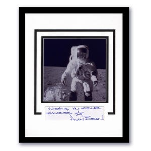 May include: Framed black and white photograph of an astronaut on the moon, wearing a spacesuit. The photograph is matted and framed in black. A handwritten note below the photo reads "Wishing you stellar success" and is signed by Alan Bean.