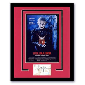 May include: Framed Hellraiser movie poster with a red mat and black frame. The poster features a character holding a puzzle box, with the text "Demon to some. Angel to others." and "HELLRAISER He'll tear your soul apart." A signed autograph is below.